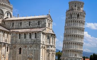 The Leaning Tower of Pisa and Pisa Cathedral with white marble facades under blue sky. The iconic tilted bell tower stands prominently beside the cathedral.