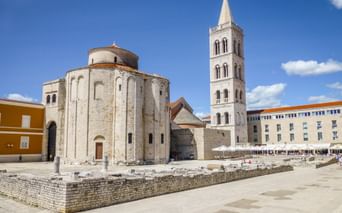 St. Donatus Church with circular stone structure and tall bell tower in Zadar's main square, with ancient Roman ruins in foreground.