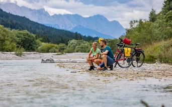 Zwei Radfahrer sitzen an einem Kiesstrand an der Isar mit beladenen Tourenrädern. Berge und Wälder im Hintergrund sichtbar.
