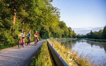 Three cyclists riding along a paved path beside the Main-Danube Canal in Bavaria, surrounded by lush green trees under a blue sky.