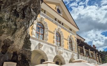 Gelbes Barockgebäude mit weißen Steinbögen und lateinischer Inschrift in Postojna. Eine große Felsformation ist links sichtbar.
