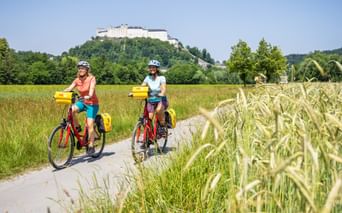 Zwei Radfahrer auf roten Rädern mit gelben Taschen fahren auf Weg durch Getreidefelder. Festung Hohensalzburg auf Hügel im Hintergrund.