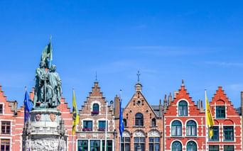 Historischer Grote Markt in Brügge mit bunten Giebelhäusern und einem Bronzedenkmal unter blauem Himmel. Fahnen schmücken die Gebäude.