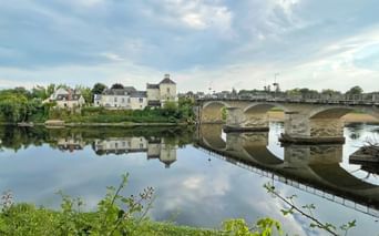 Mehrbogige Steinbrücke über die Loire in Chinon, mit historischen Gebäuden, die sich im ruhigen Wasser spiegeln, und grüner Vegetation im Vordergrund.