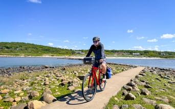 Cyclist on red bike riding along narrow coastal path surrounded by rocks and green vegetation, with blue water and coastline in background.