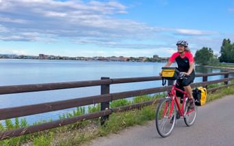 Radfahrer mit rotem Fahrrad und gelben Taschen auf Uferweg in Friaul, mit Holzgeländer, ruhiger Lagune und Küstenstadt im Hintergrund.