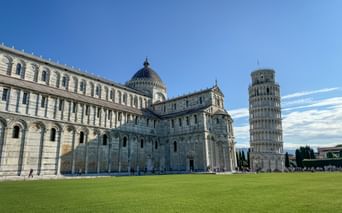 Pisa Cathedral (Santa Maria Assunta) with its striped marble facade and the famous Leaning Tower on green lawn under blue sky.
