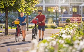 Zwei Radfahrer schieben ihre Räder durch einen sonnigen Park in Bad Ischl. Weiße Blumen im Vordergrund, Kongresshaus im Hintergrund sichtbar.