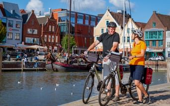 Zwei Radfahrer mit Rädern am Husumer Hafen. Bunte historische Häuser und Boote säumen die Uferpromenade unter blauem Himmel.