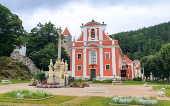 Orange Barockkirche Maria Heimsuchung mit weißen Verzierungen in Böhmen, umgeben von grünen Rasenflächen, Blumenbeeten und bewaldeten Hügeln.