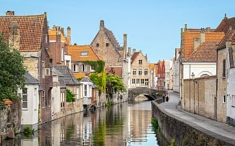Picturesque canal in Bruges with medieval brick houses, orange-tiled roofs, and a stone bridge. Trees line the waterway under a clear blue sky.