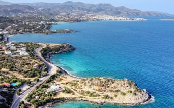 Luftaufnahme der Küste Kretas mit kurvenreicher Straße entlang türkiser Buchten, felsigen Halbinseln und Bergen im Hintergrund.