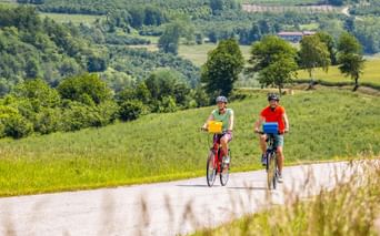 Zwei Radfahrer auf einer Landstraße durch grüne Hügel in Slowenien. Einer trägt ein grünes Shirt, der andere rot, mit üppiger Tallandschaft.