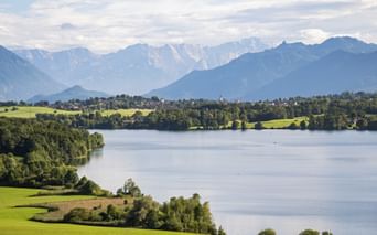 View of Riegsee lake surrounded by green meadows and forests with snow-capped Alpine mountains in the background under a cloudy sky.