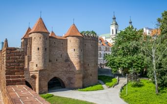 Warschauer Barbakan-Festung mit roten Ziegeltürmen und Kegeldächern. Grüne Rasenhänge und eine Kirche mit weißen Türmen im Hintergrund.