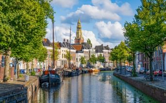Gracht in Groningen mit historischen Booten, bunten Häusern und hohem Kirchturm unter blauem Himmel mit weißen Wolken.