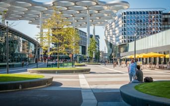 Utrechter Bahnhofsvorplatz mit markanter weißer Überdachung und geschwungenem modernen Gebäude. Menschen entspannen auf Bänken nahe Bäumen.