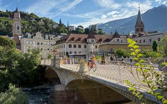 Radfahrer überqueren eine verzierte Steinbrücke über einen Fluss in Meran, mit historischen Gebäuden, Kirchtürmen und Bergen im Hintergrund.