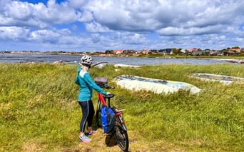Cyclist with bike on grassy coastal area overlooking water and village houses in Träslövsläge, Sweden. Blue sky with white clouds above.