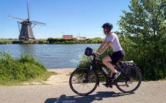 Female cyclist on path beside canal with traditional Dutch windmill in Kinderdijk under blue sky.