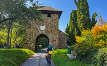 Radfahrer mit gelben Taschen fährt durch gewölbtes Backsteintor am Murradweg. Grüne Wiese, Zypressen und Kirchturm im Hintergrund sichtbar.