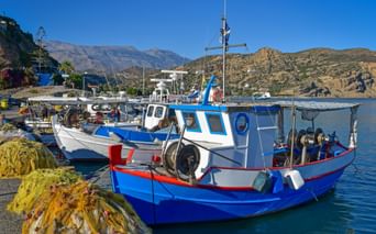 Blau-weiße Fischerboote im kretischen Hafen mit gelben Netzen am Kai. Berge und klarer blauer Himmel im Hintergrund sichtbar.