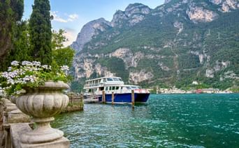 Blue and white ferry docked at Riva del Garda pier with turquoise Lake Garda waters and dramatic mountain cliffs in the background.