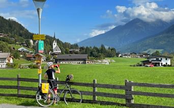 Radfahrer mit gelben Taschen trinkt an Brunnen neben Holzzaun. Alpendorf mit Kirche und Bergen im Hintergrund unter blauem Himmel.