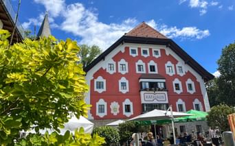 Red and white Rathaus building in Saalfelden with outdoor café seating under green umbrellas, framed by bright green foliage.
