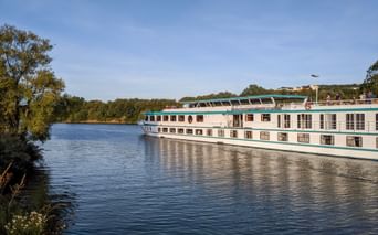 MS Switzerland Flusskreuzfahrtschiff mit weißem Rumpf und türkisfarbenen Akzenten auf ruhigem Fluss, umgeben von grünen Bäumen.