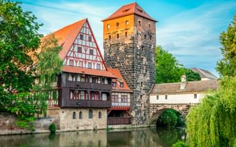 Medieval stone tower and half-timbered house beside river in Nuremberg, with stone bridge and weeping willows under blue sky.