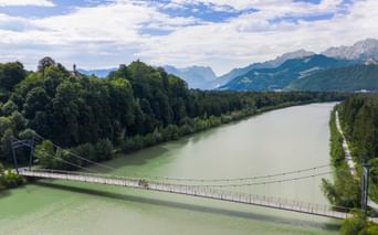 Suspension bridge crossing green river with cyclists, surrounded by forests and Alpine mountains under blue sky near Puch on Alpe-Adria route.