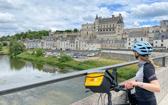 Radfahrerin mit blauem Helm auf Brücke mit Blick auf die Loire und Schloss Amboise. Das Renaissance-Schloss thront über historischen Gebäuden.