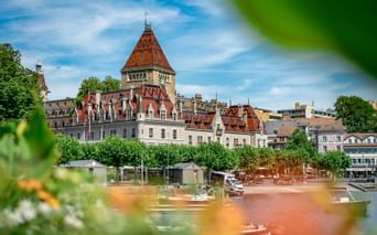 Schloss Château d'Ouchy mit rotem Ziegeldach und Turm in Lausanne am Genfersee, eingerahmt von grünem Laub und bunten Blumen im Vordergrund.