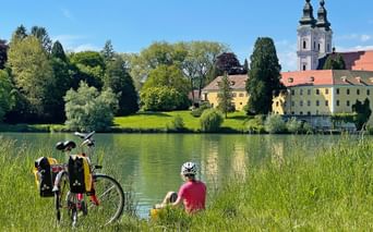 Radfahrer in rotem Shirt und Helm sitzt am See mit Fahrrad. Gelbe Klostergebäude und Kirche mit Doppeltürmen am gegenüberliegenden Ufer.
