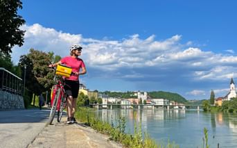 Cyclist in pink shirt with yellow bag on Danube riverside path in Passau, with river, historic buildings and church visible across the water.