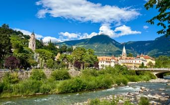 Die Passer fließt durch Meran mit historischen Gebäuden, Kirchtürmen und Bergen in Südtirol unter blauem Himmel mit weißen Wolken.