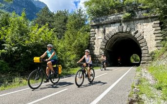 Family cyclists on Alpe Adria route approaching historic stone tunnel near Dogna, surrounded by green mountains and trees.