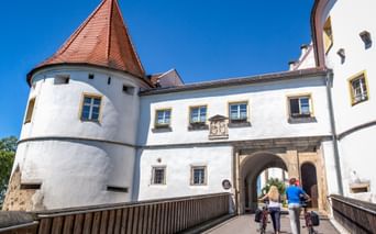 Zwei Radfahrer mit Rädern an der Eingangsbrücke von Schloss Wörth, einem weißen Gebäude mit rotem Dach und Rundturm unter blauem Himmel.