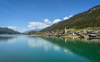 Turquoise Weissensee lake with Techendorf village on the shore, featuring a church spire and traditional houses against forested mountains.
