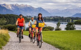 Three cyclists with helmets riding along Riegsee lake in Bavaria. Mountains and green meadows visible in background under cloudy sky.