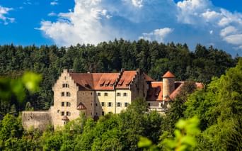 Burg Rabenstein mit roten Ziegeldächern und rundem Turm, umgeben von grünem Wald unter dramatischen weißen Wolken und blauem Himmel in Österreich.
