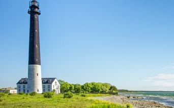 Tall black and white lighthouse with keeper's house on Baltic Sea coast. Green meadow in foreground, rocky beach and blue sky visible.