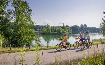 Three cyclists riding along a paved path beside a calm green river with trees and forest in the background under a blue sky.