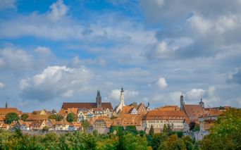 Panoramic view of Rothenburg ob der Tauber with medieval buildings, orange-tiled roofs, and church towers above green trees under a cloudy sky.