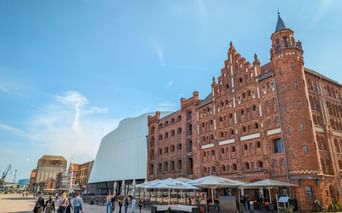 Red brick Türmchenspeicher building with tower in Stralsund, adjacent to modern white structure. People gather on the plaza under blue sky.