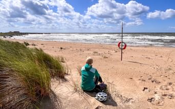 Radfahrer in türkisfarbenem Shirt sitzt am Sandstrand mit Helm neben sich und blickt aufs Meer. Dünengras links, Rettungsring am Strand.