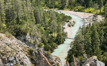 Aerial view of turquoise Isar River winding through dense forest with rocky banks and a small footbridge crossing the water.