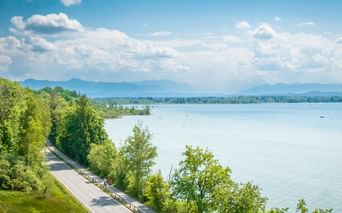 Scenic cycling path along Lake Starnberg with lush green trees, calm blue water, and mountain ranges in the distance under a cloudy sky.