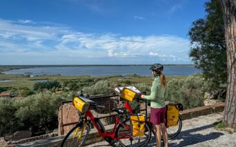 Radfahrerin mit rotem Tourenrad und gelben Packtaschen mit Blick auf den Lago di Massaciuccoli in der Toskana unter blauem Himmel.